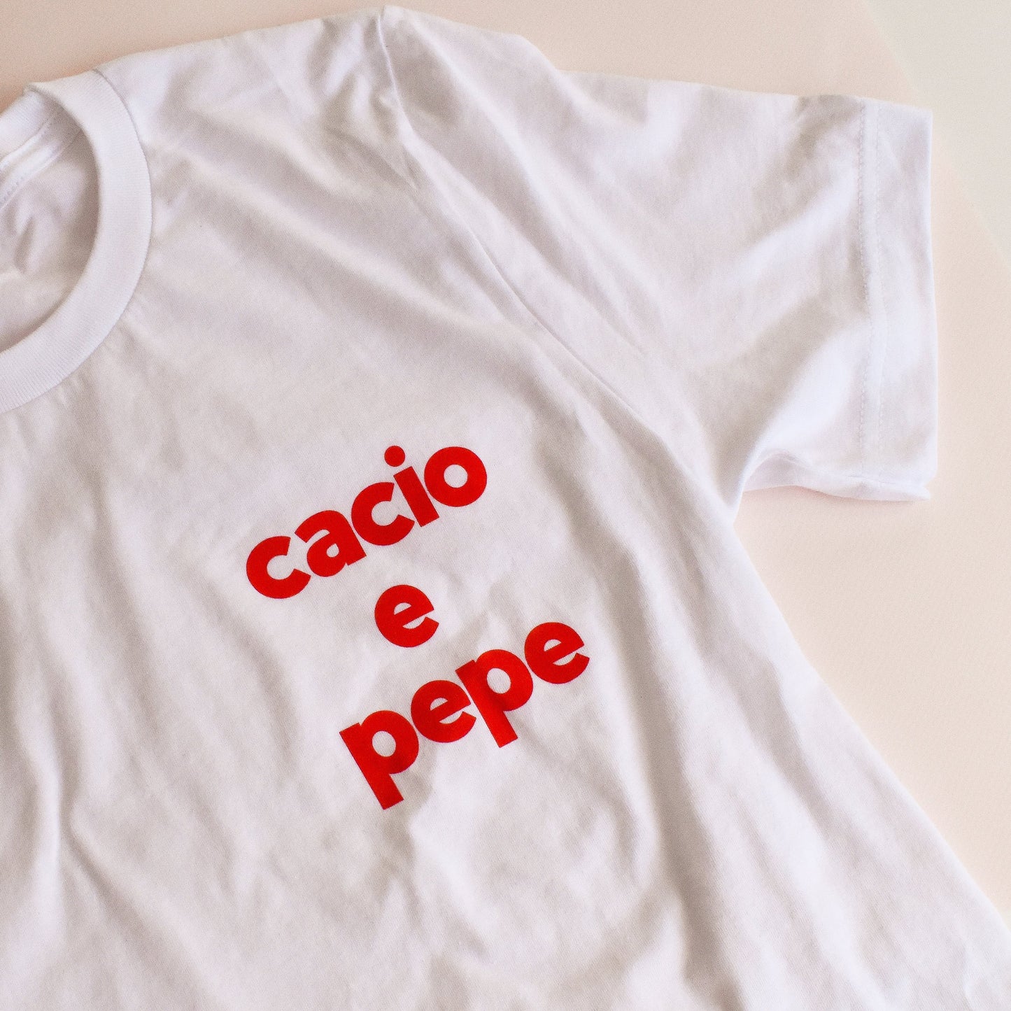 Image of a white t-shirt on a pale pink background. The t-shirt features a bold, red text that reads Cacio E Pepe, on the left side of the chest. 